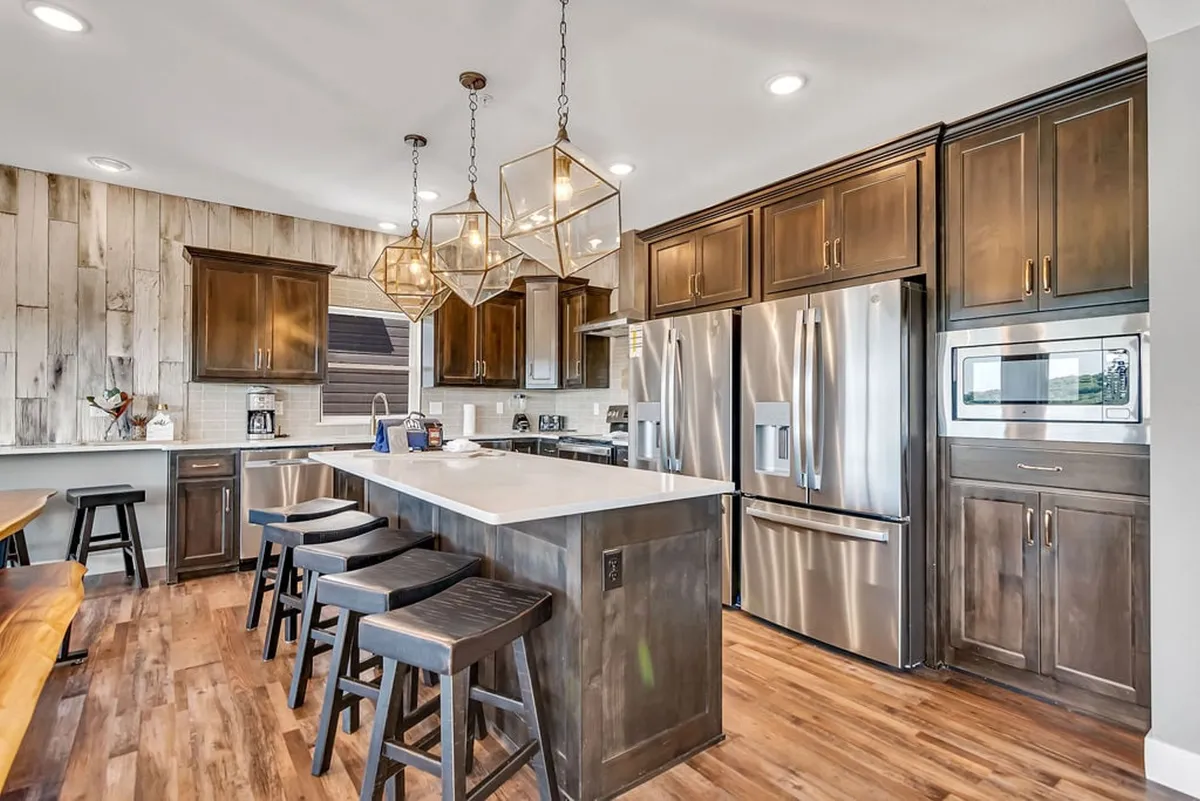 A spacious kitchen with a prominent island, hanging light fixtures, stainless steel appliances, and warm wooden accents, surrounded by ample seating. The space is set up for both cooking and casual gathering, ideal for group meals and celebratory moments.