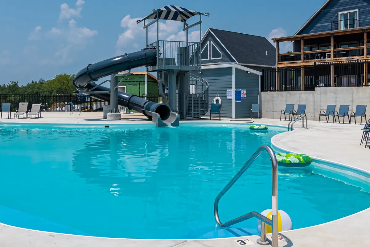 A lively resort pool area featuring a swimming pool with a water slide, lounge chairs, and colorful inflatable rings. Kids are seen playing on the slide while adults relax on the deck, all framed by a welcoming, sunny setting.