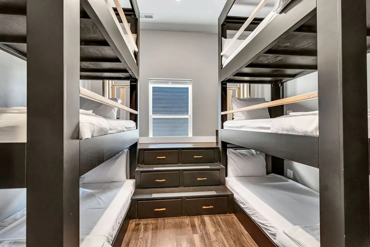 A custom-designed bunk room with built-in bunk beds, integrated storage drawers, and a window letting in natural light. The setup is playful and inviting, clearly oriented toward children, with enough space for sleepovers, story time, and play.