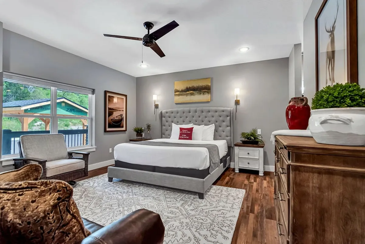 A spacious king suite with a king-sized bed featuring a tufted headboard, large window offering natural light and scenic views, a stylish area rug, elegant wooden dresser, and tasteful artwork on the wall. The overall mood is cozy and welcoming, with plush bedding and a relaxing atmosphere that invites rest and comfort.