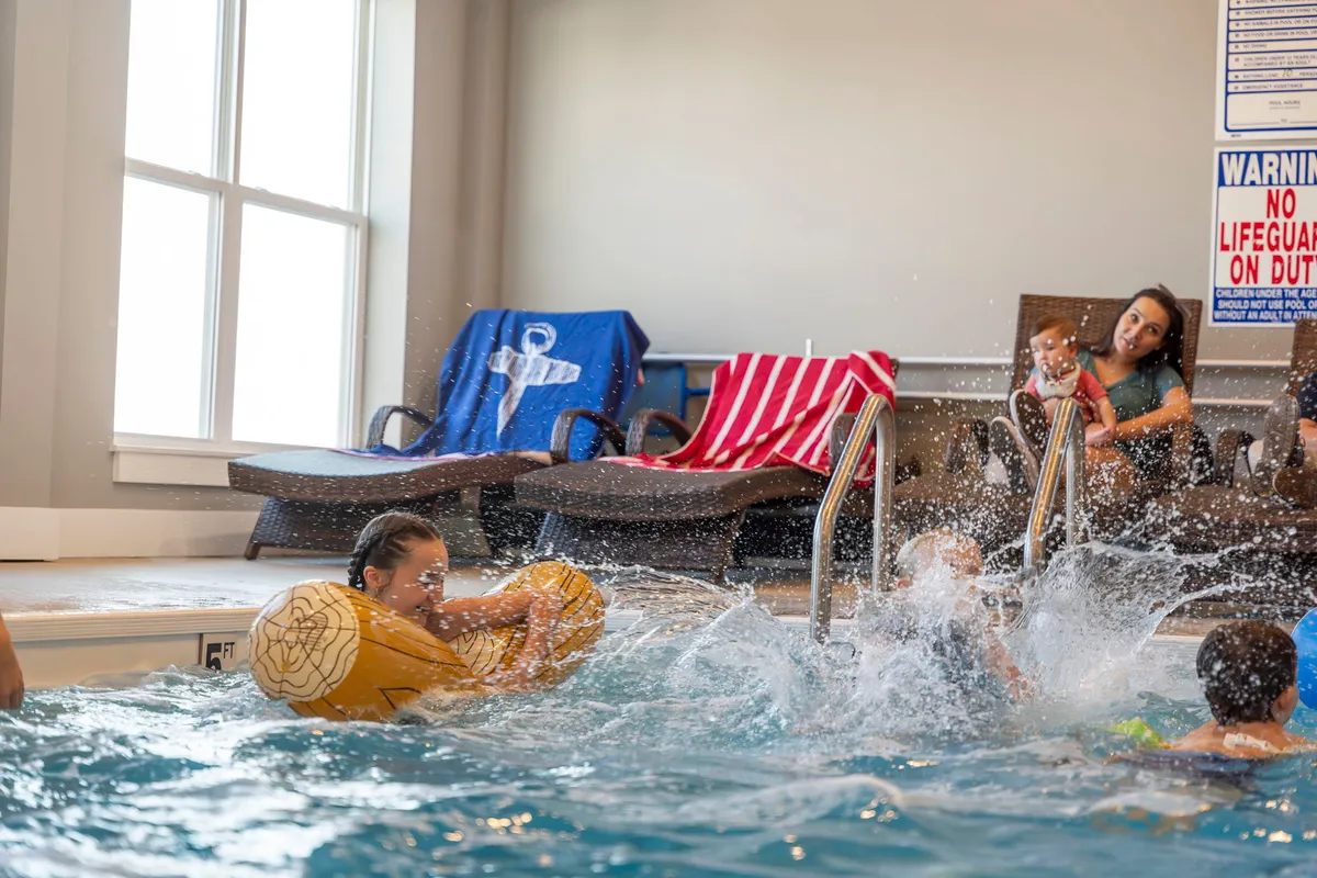 A lively indoor pool scene with children joyfully splashing in the water while an adult supervises from the side. Comfortable poolside chairs line the edges, creating a relaxed, family-friendly atmosphere filled with laughter and fun.
