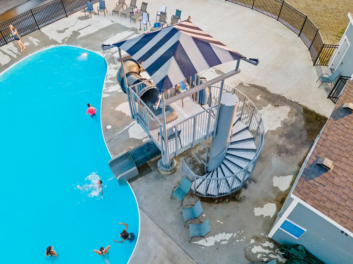This exterior image features a vibrant outdoor swimming pool with a water slide, sun loungers, and children swimming and playing. The scene is lively and full of energy, suggesting summer fun and family activity. The surrounding pool area is set for socializing and sunbathing, capturing the essence of warm-weather relaxation and adventure.
