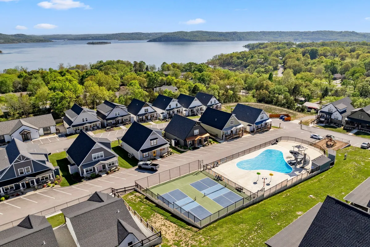 An inviting exterior view showcasing a large swimming pool, tennis courts, and a beautiful lake beyond, nestled among neighborhood houses. The recreational amenities and the scenic backdrop evoke outdoor activity, relaxation, and easy access to resort fun.