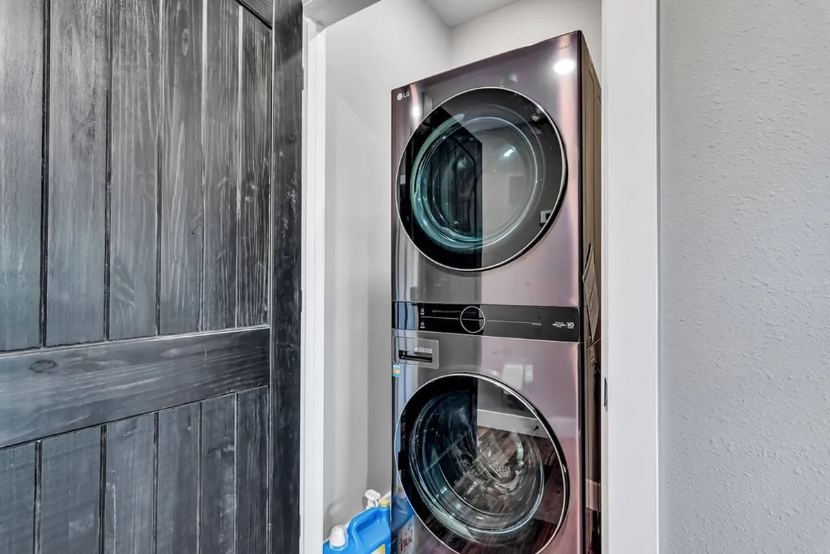 A modern laundry area featuring stacked washer and dryer units beside a dark wooden door, with shelves neatly stocked with laundry supplies. The space is compact, efficient, and clearly designed for guest convenience.