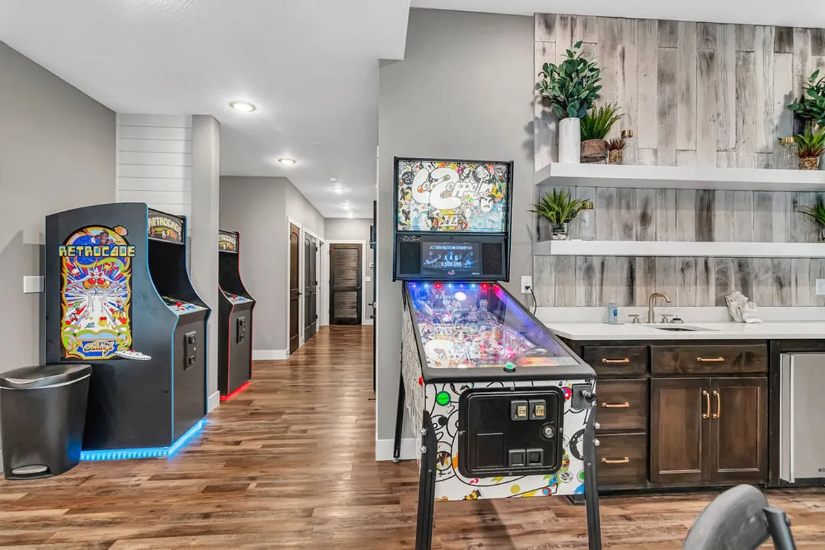 A vibrant game room featuring several stand-up arcade machines, a pinball machine, open shelving with decorative plants, and space to mingle. The setting is lively and colorful, capturing the excitement of tournaments and nostalgic gaming nights.