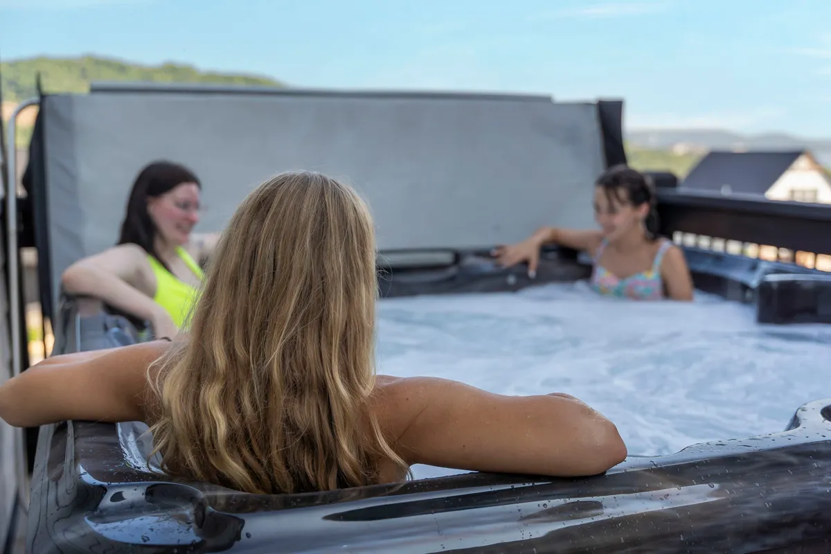 A group of women enjoy a relaxing soak in a private outdoor hot tub, surrounded by a scenic landscape of hills and open skies. The mood is cheerful, and the setting highlights both exclusivity and the opportunity to unwind while taking in stunning views.