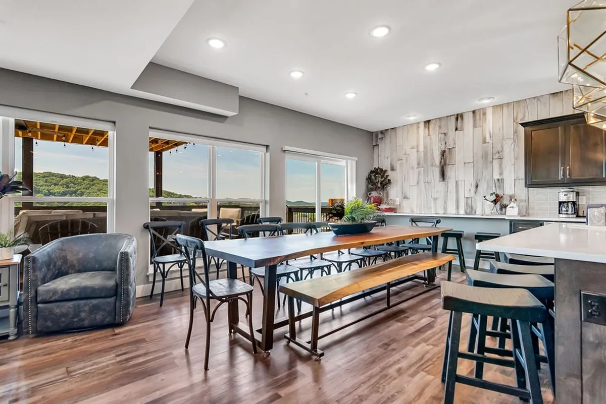 A warm, farmhouse-inspired great room featuring a large dining table paired with a spacious kitchen island, all set against a backdrop of panoramic windows framing stunning lake views. The wood accent wall and stylish lighting create a welcoming atmosphere perfect for family gatherings and celebrations.