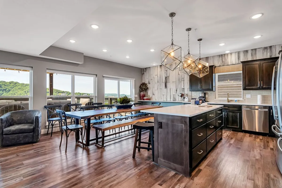 A spacious, modern kitchen and dining area with a central island, large dining table, contemporary light fixtures, and windows offering scenic views. The space radiates hospitality, ideal for group gatherings, shared meals, and spring or autumn celebrations.