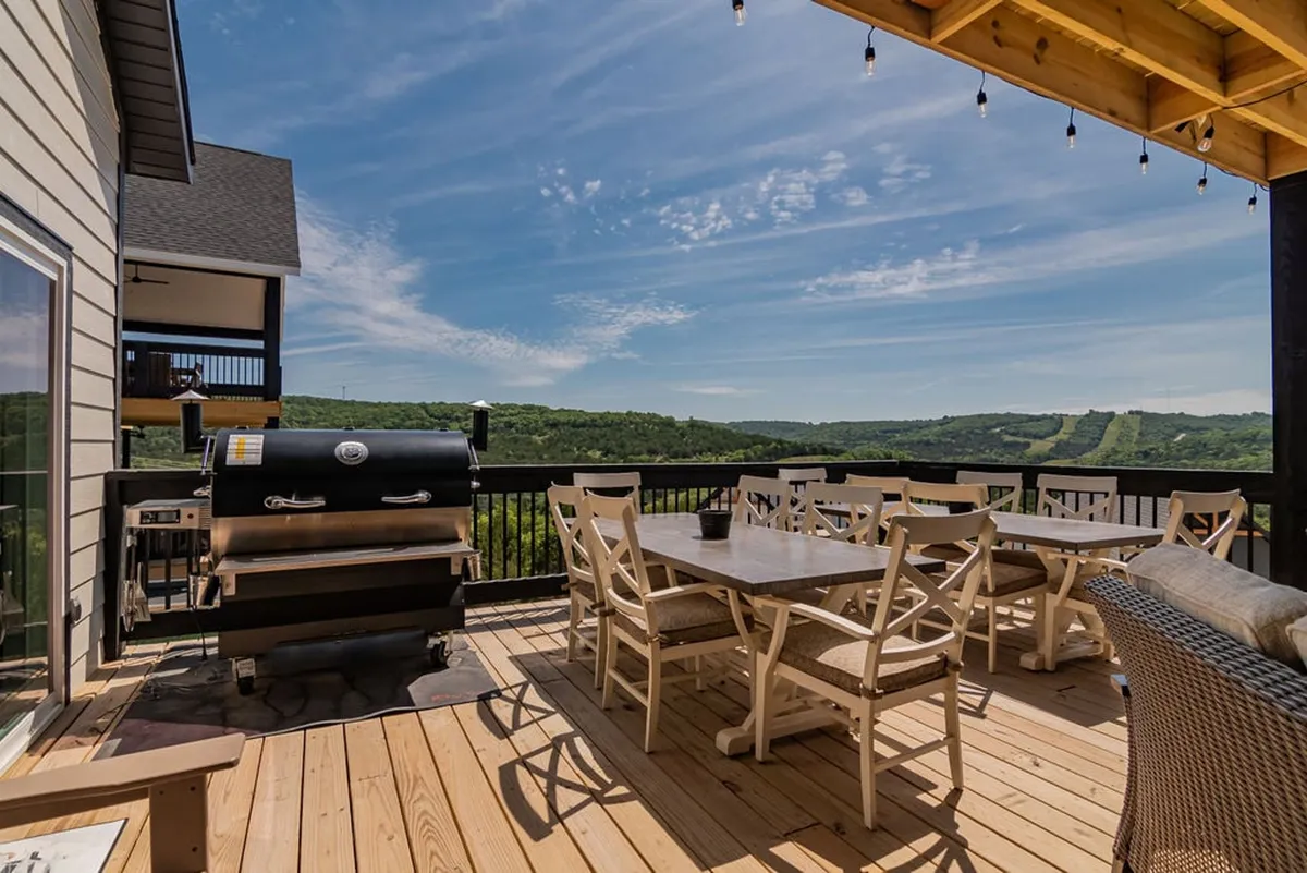 A welcoming outdoor dining area featuring a large table set under string lights, a modern grill nearby, and expansive views of rolling hills and greenery. The scene is festive and ready for a barbecue or group meal, inviting guests to gather and enjoy the outdoors together.